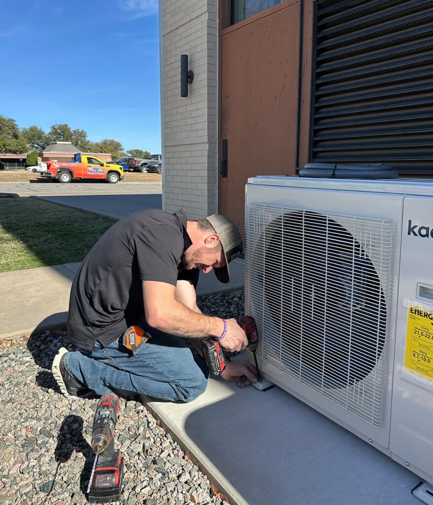 HVAC technician kneeling to install mini split condenser unit with power drill