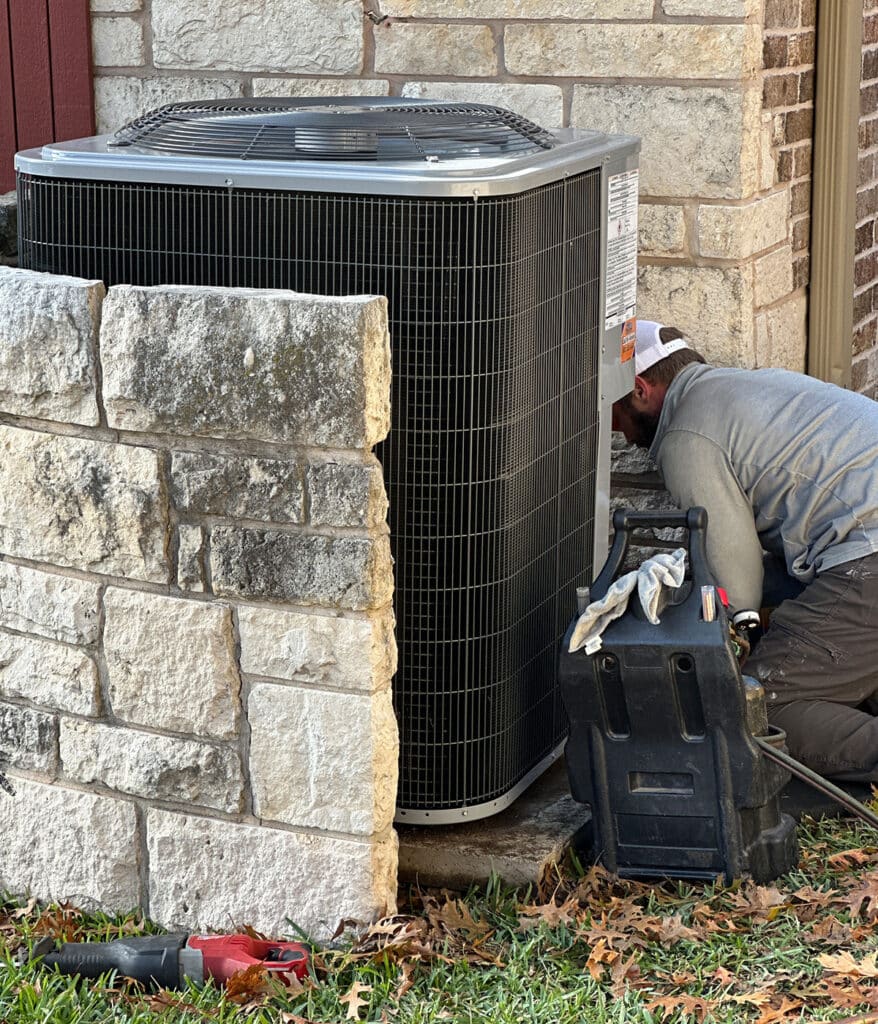 HVAC technician servicing air conditioner condenser unit beside stone wall with recovery machine